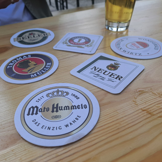 A collection of beer mats with various designs featuring famous German football players placed on a wooden table with a glass of beer.