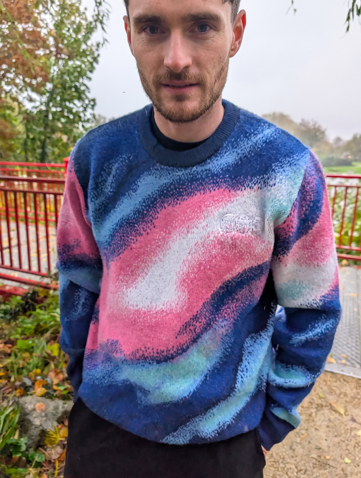 A man wearing a fuzzy crewneck sweatshirt with a pink, blue, and white abstract swirl pattern, standing outdoors in front of a red railing and trees.